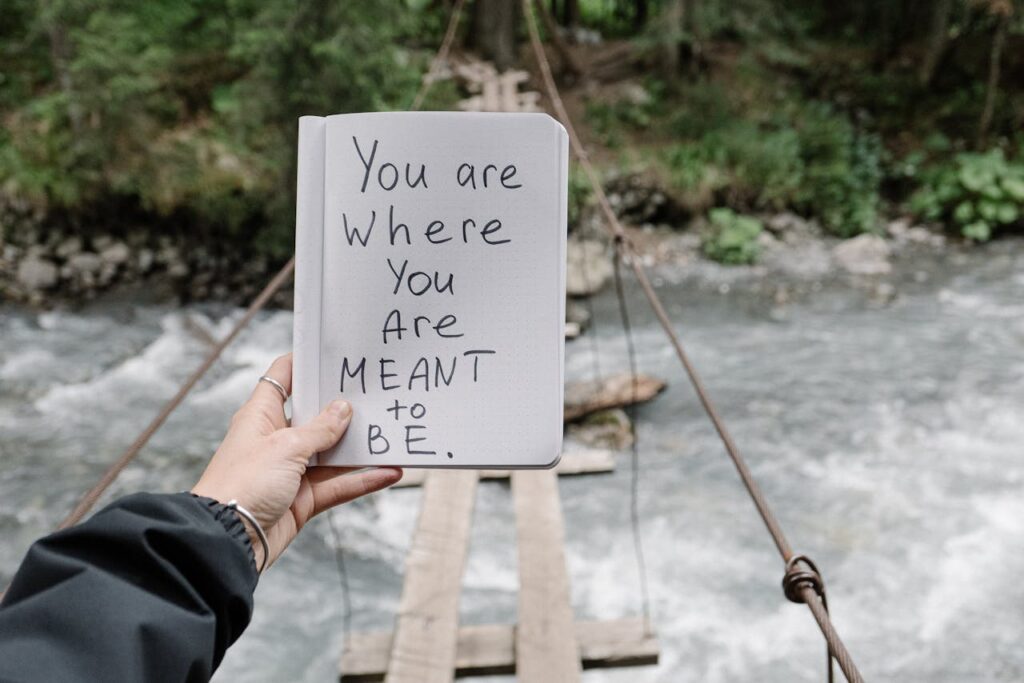 Close-up of a notebook with an inspiring message held over a river with a wooden bridge.